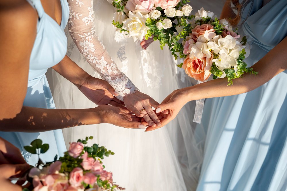 Bride showing her wedding ring with bridesmaids in light blue dresses holding floral bouquets.